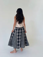 A woman viewed from behind wearing a sleeveless cream top and a black and white plaid midi skirt, standing barefoot against a plain off-white studio background.