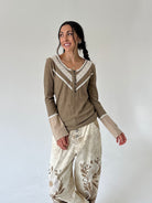 A woman modeling an olive green long-sleeve henley top with cream lace trim and cream harem-style pants with brown floral embroidery, set against a plain light gray studio background.