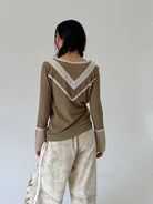 A woman shown from the back wearing an olive green long-sleeve top with cream lace trim and cream wide-leg pants, set against a plain light gray studio background.