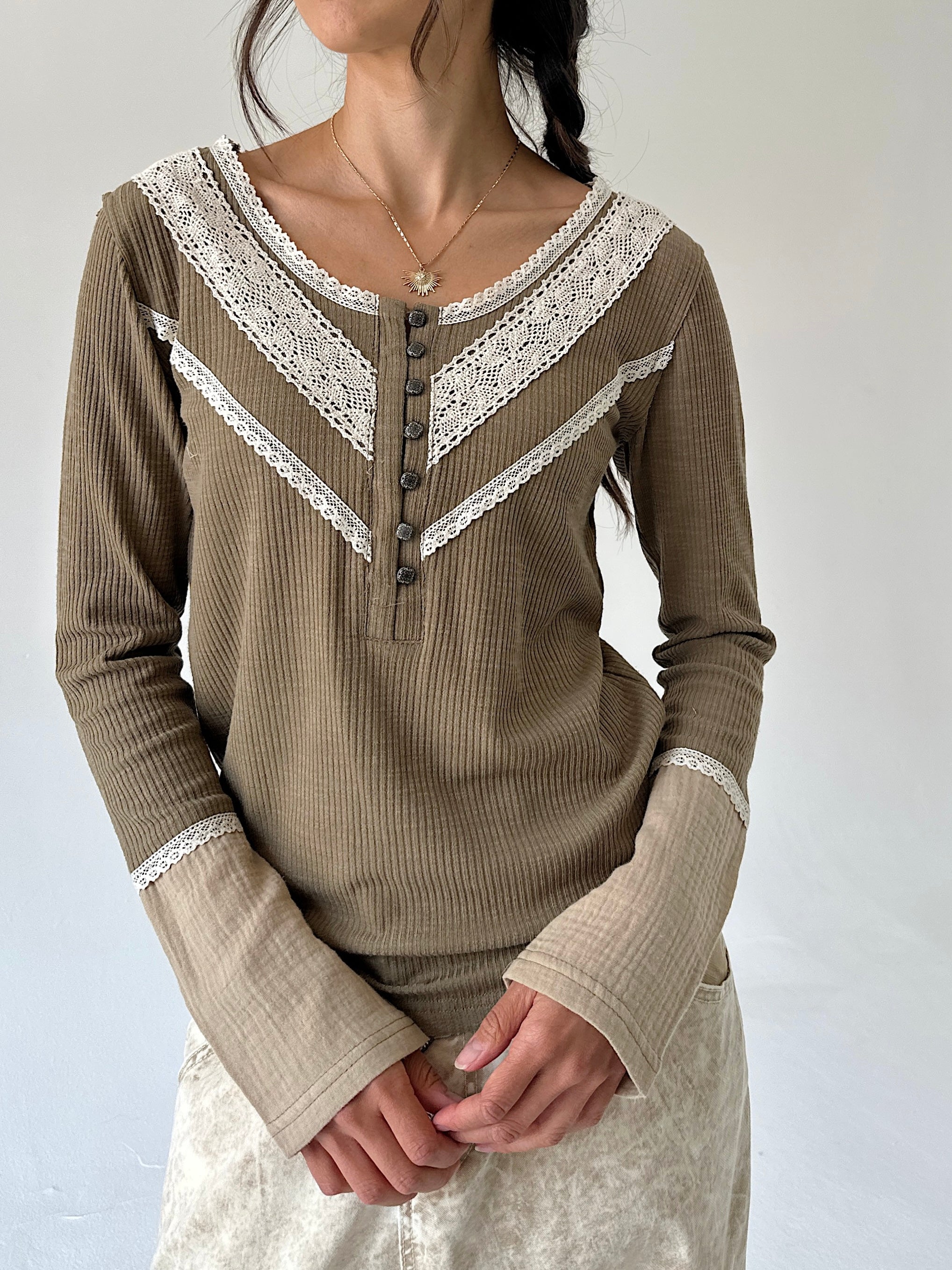A woman wearing a ribbed long-sleeve henley top in taupe olive with white lace trim and button details, paired with a light patterned skirt against a plain light gray background.