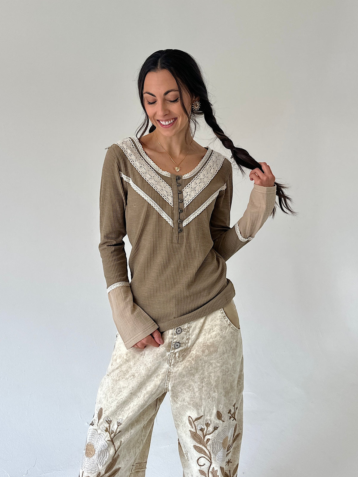 A woman modeling an olive long-sleeve henley top with cream lace trim and matching cream pants against a plain light gray studio background.