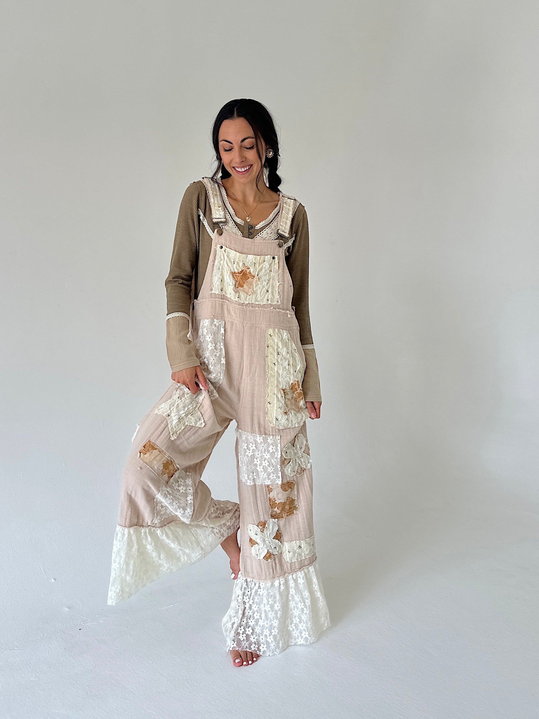 A woman modeling a bohemian-style patchwork jumpsuit in beige and brown, featuring floral lace panels and wide-leg flared pants, against a plain light gray studio background.