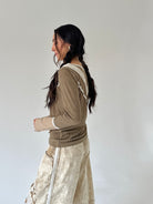 A woman with long dark hair in a side braid is shown from the side/back wearing a brown olive ribbed long-sleeve top with cream lace trim and a cream textured skirt, against a plain light gray studio background.