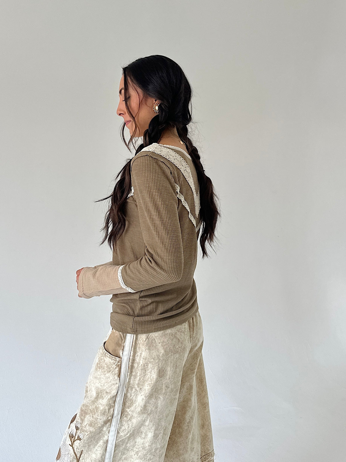 A woman with long dark hair in a side braid is shown from the side/back wearing a brown olive ribbed long-sleeve top with cream lace trim and a cream textured skirt, against a plain light gray studio background.