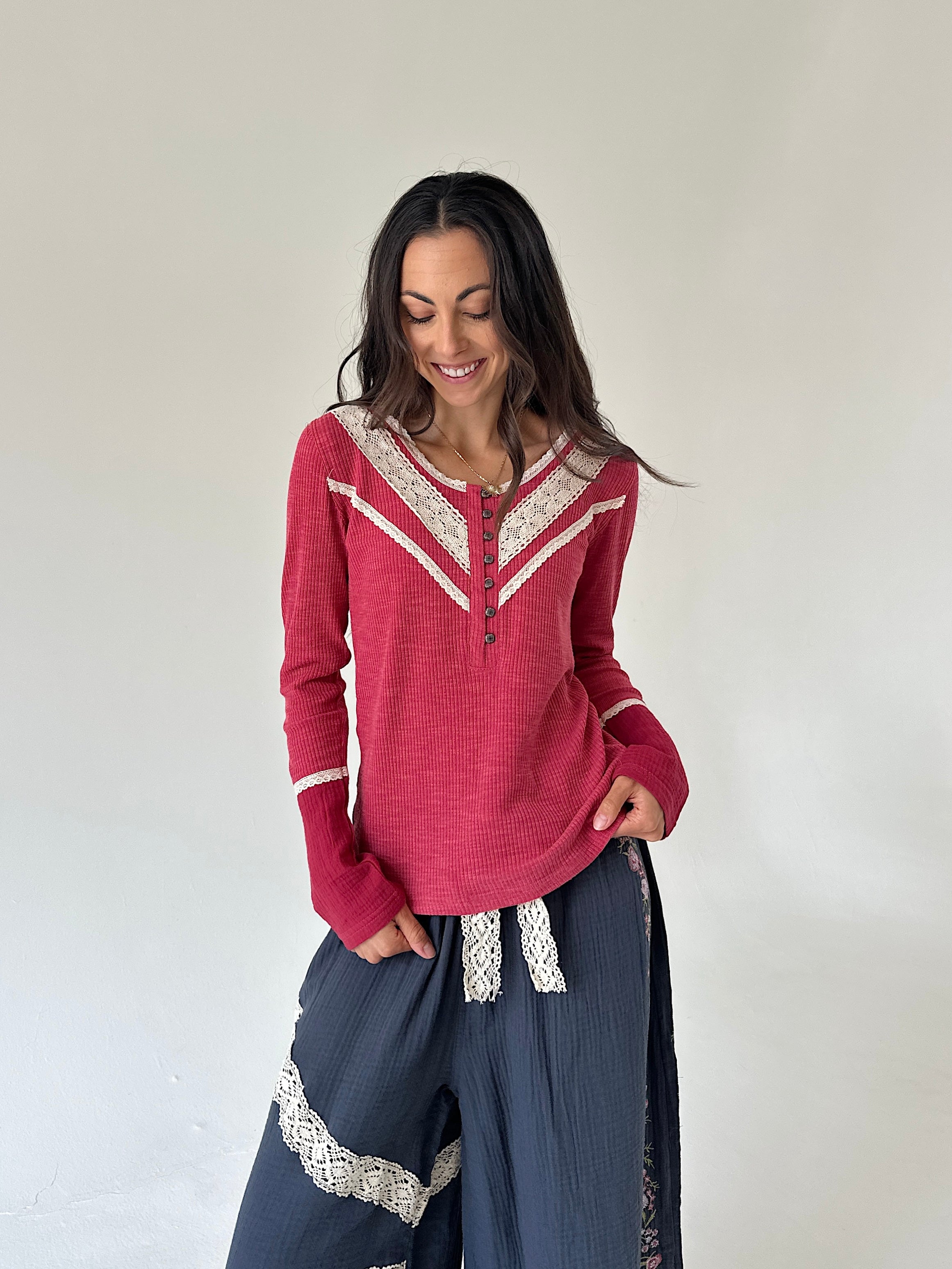 A woman modeling a long-sleeve henley top in dusty rose pink with cream lace trim, paired with dark blue wide-leg pants with lace details, against a plain off-white wall.