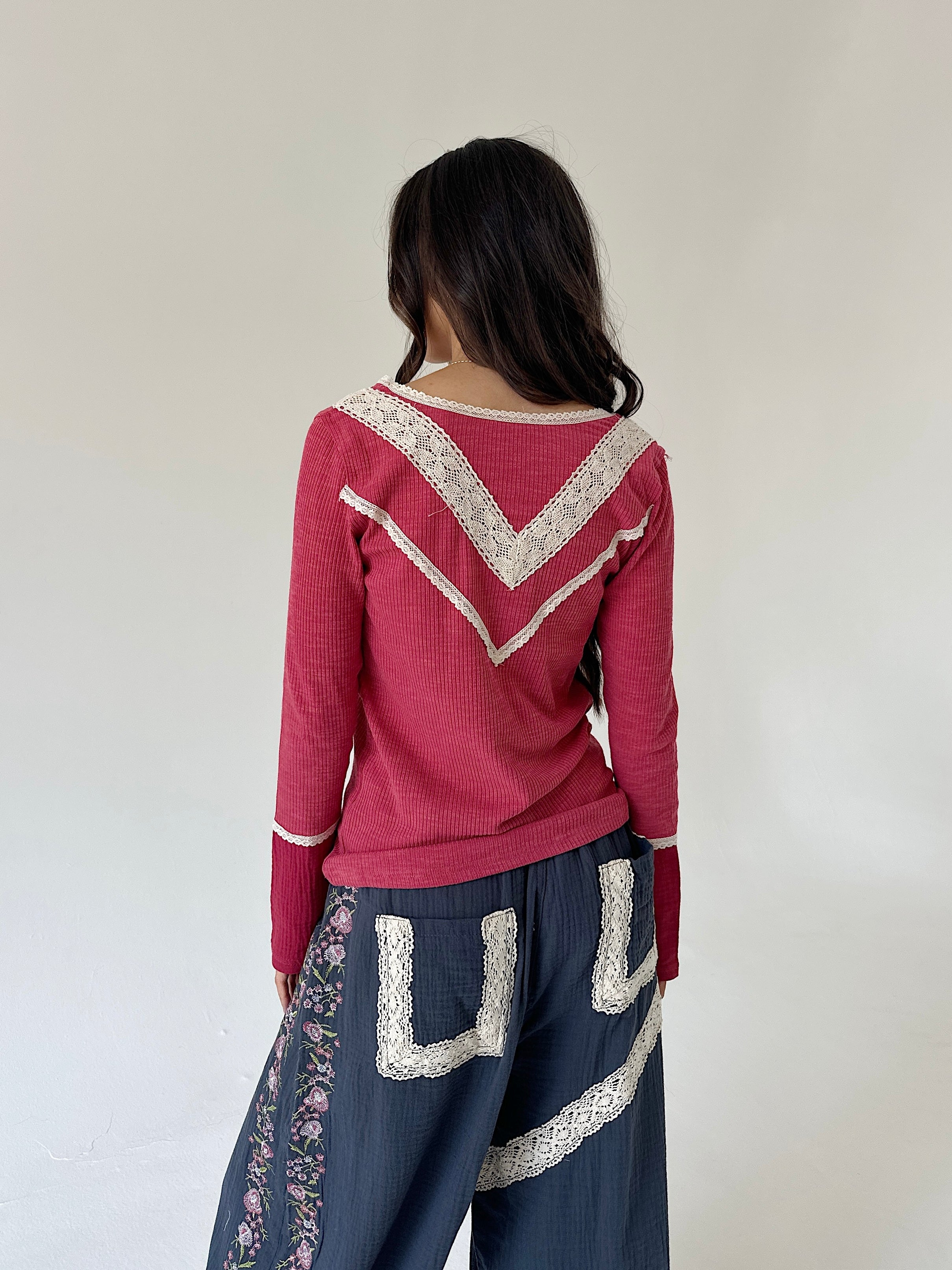 A woman shown from behind wearing a dusty rose long-sleeve knit top with cream lace trim and dark blue wide-leg pants with floral and lace details, set against a plain off-white wall.