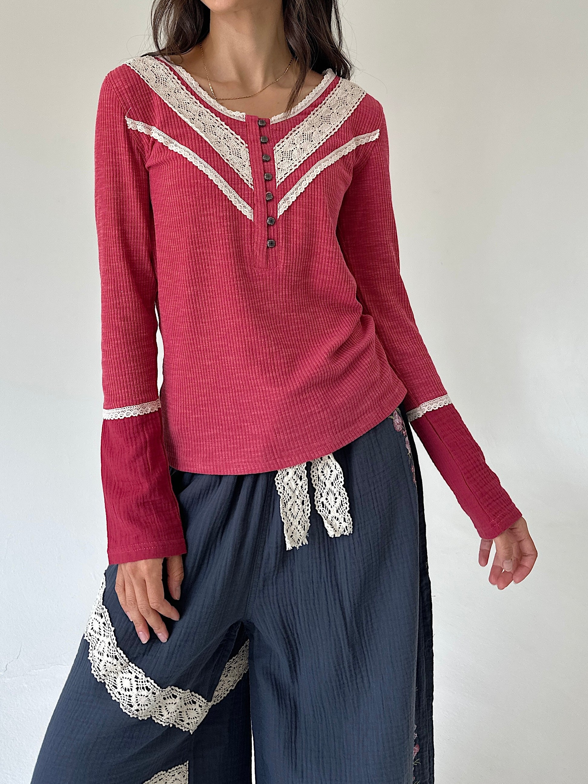 A woman wearing a long-sleeve knit top in dusty rose pink with cream lace trim along the V-neckline and cuffs, paired with dark blue wide-leg pants featuring cream lace details.