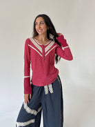 A woman modeling a long-sleeve knit top in dusty rose pink with cream lace trim, paired with dark blue gauze pants featuring lace accents, against a plain off-white wall.