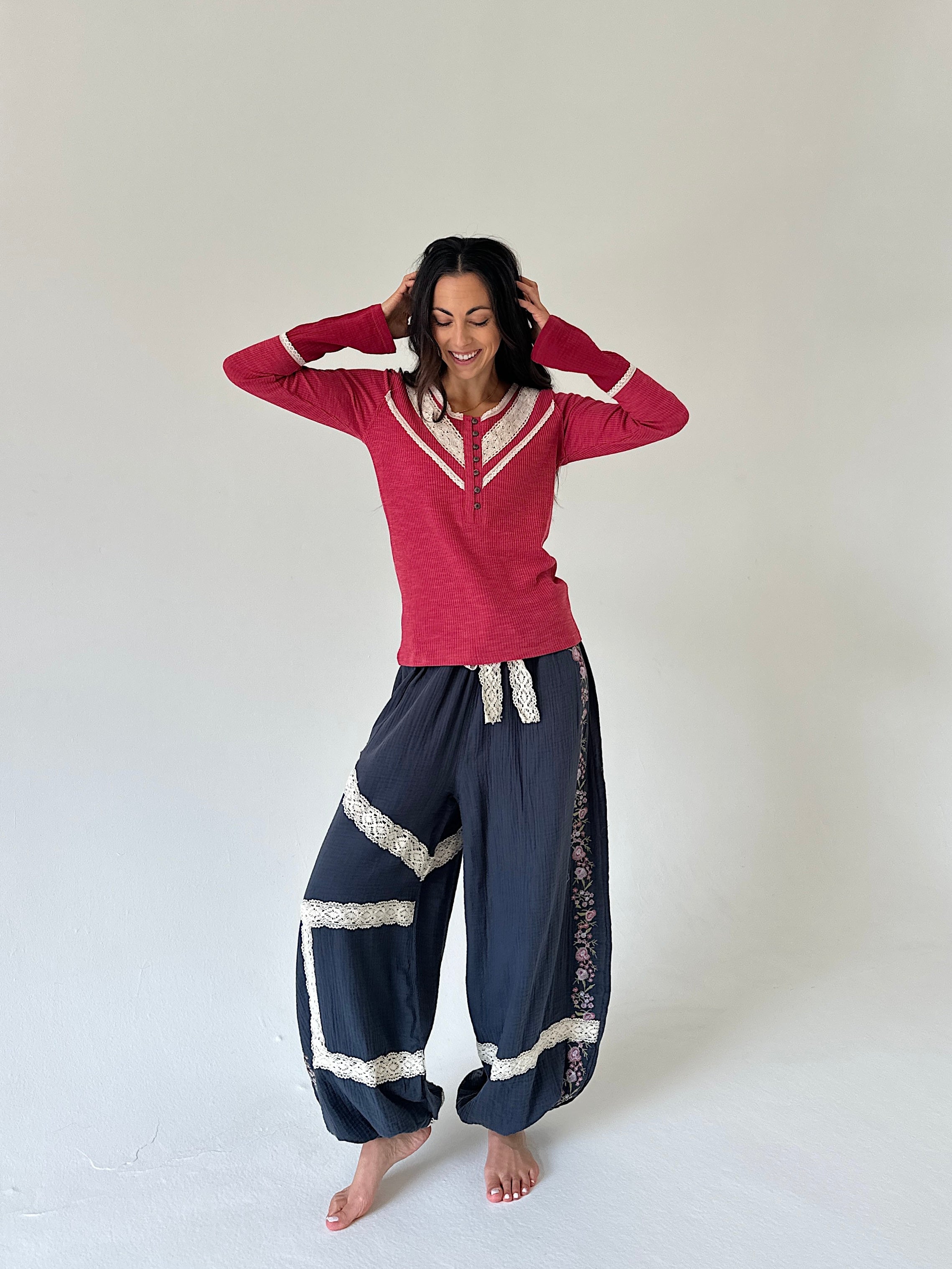 A woman modeling a red long-sleeve henley shirt with white trim and navy blue harem pants with white lace details, standing against a plain light gray studio background.