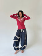 A woman modeling a red long-sleeve henley shirt with white trim and navy blue harem pants with white lace details, standing against a plain light gray studio background.
