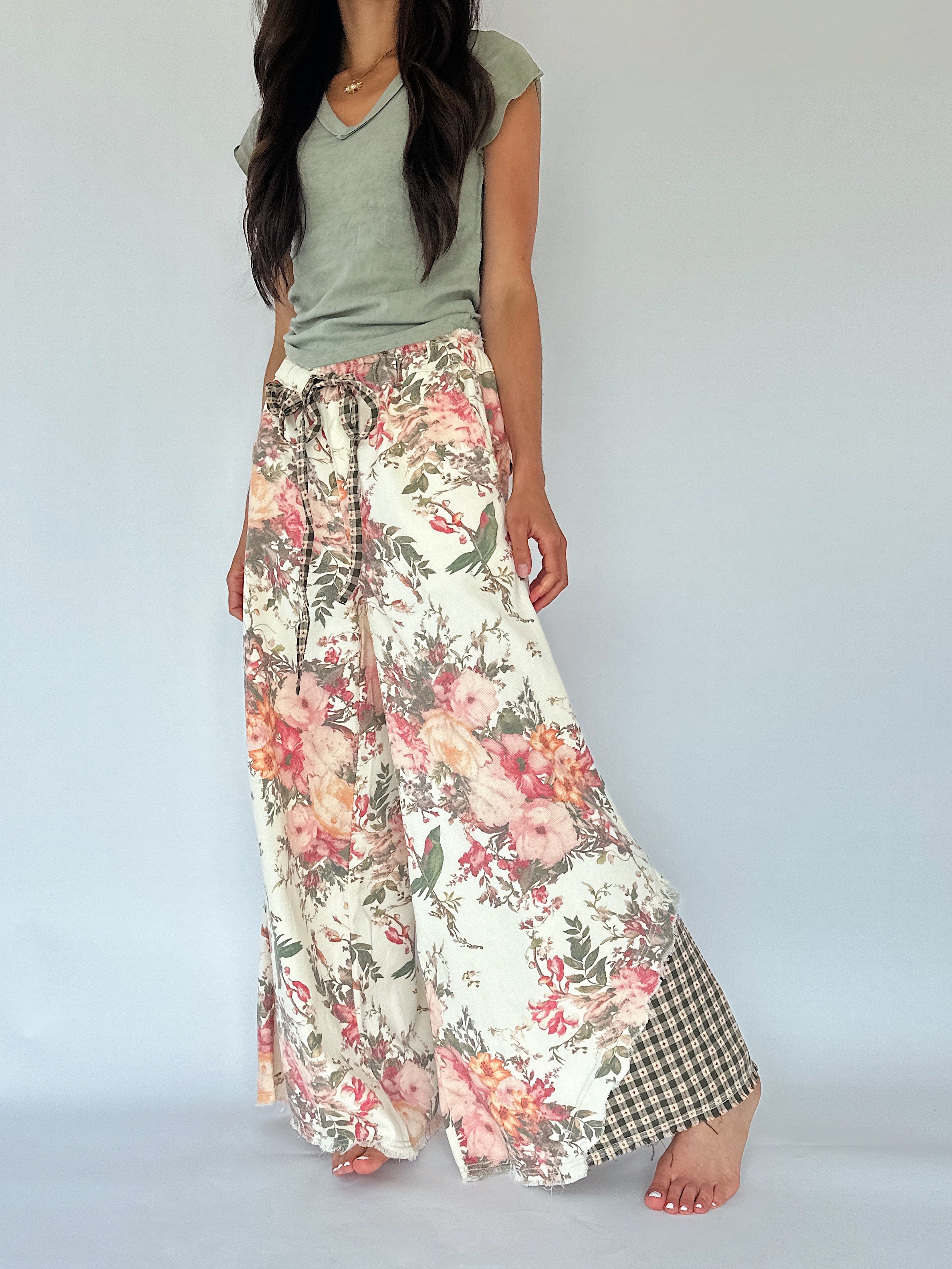 A woman modeling wide-leg floral pants with a cream base, pink and red flowers, green leaves, and a green-and-white gingham check panel at the hem, paired with an olive green top against a light gray studio background.