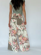 A person shown from the back wearing wide-leg floral pants with gingham patch pockets and a sage green top, photographed against a plain light gray studio background.