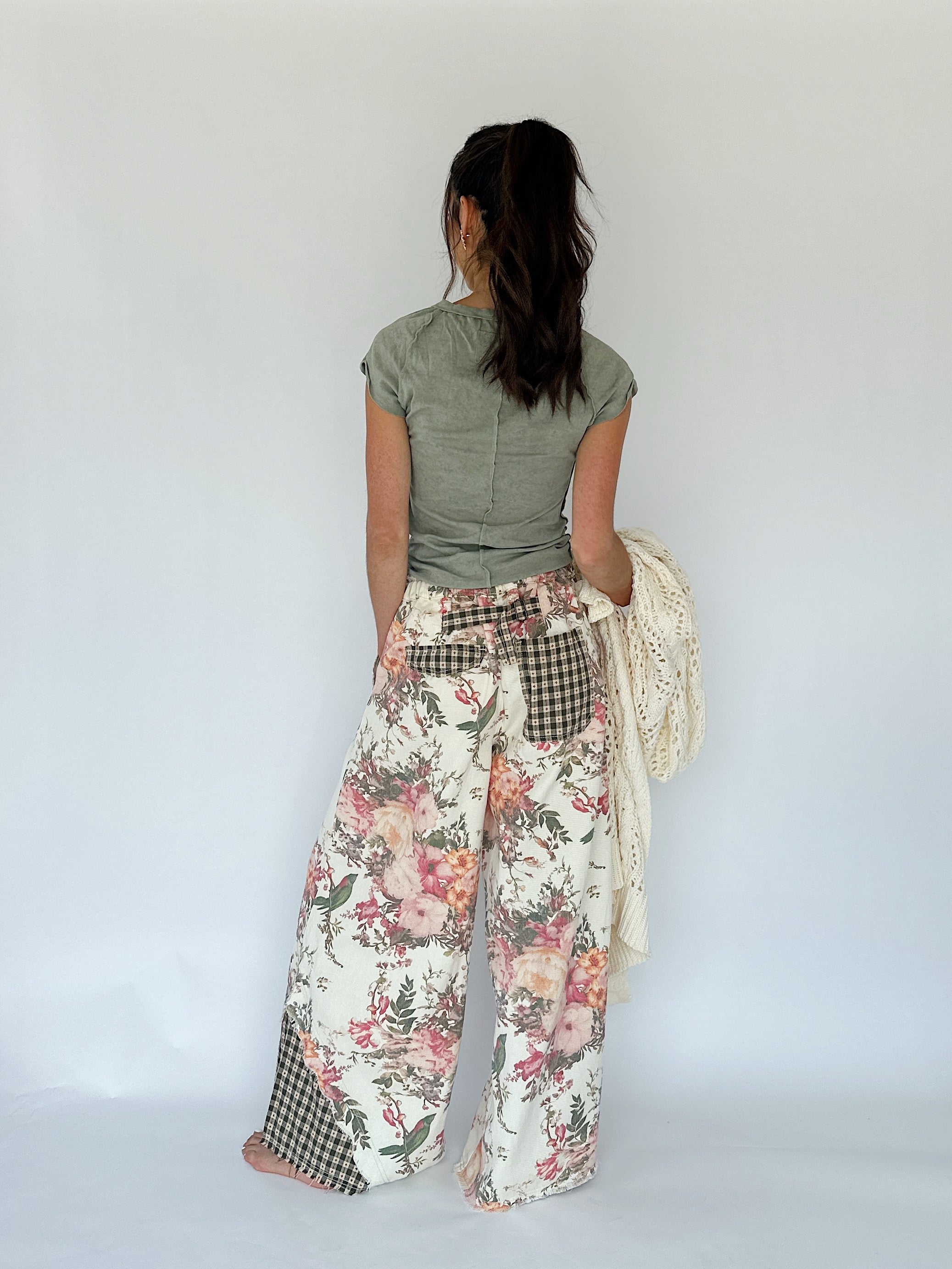 A woman shown from behind wearing wide-leg patchwork pants with a floral print, gingham pockets, and a green top, holding a cream knit sweater against a plain white studio background.