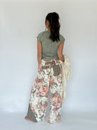 A woman shown from behind wearing wide-leg patchwork pants with a floral print, gingham pockets, and a green top, holding a cream knit sweater against a plain white studio background.