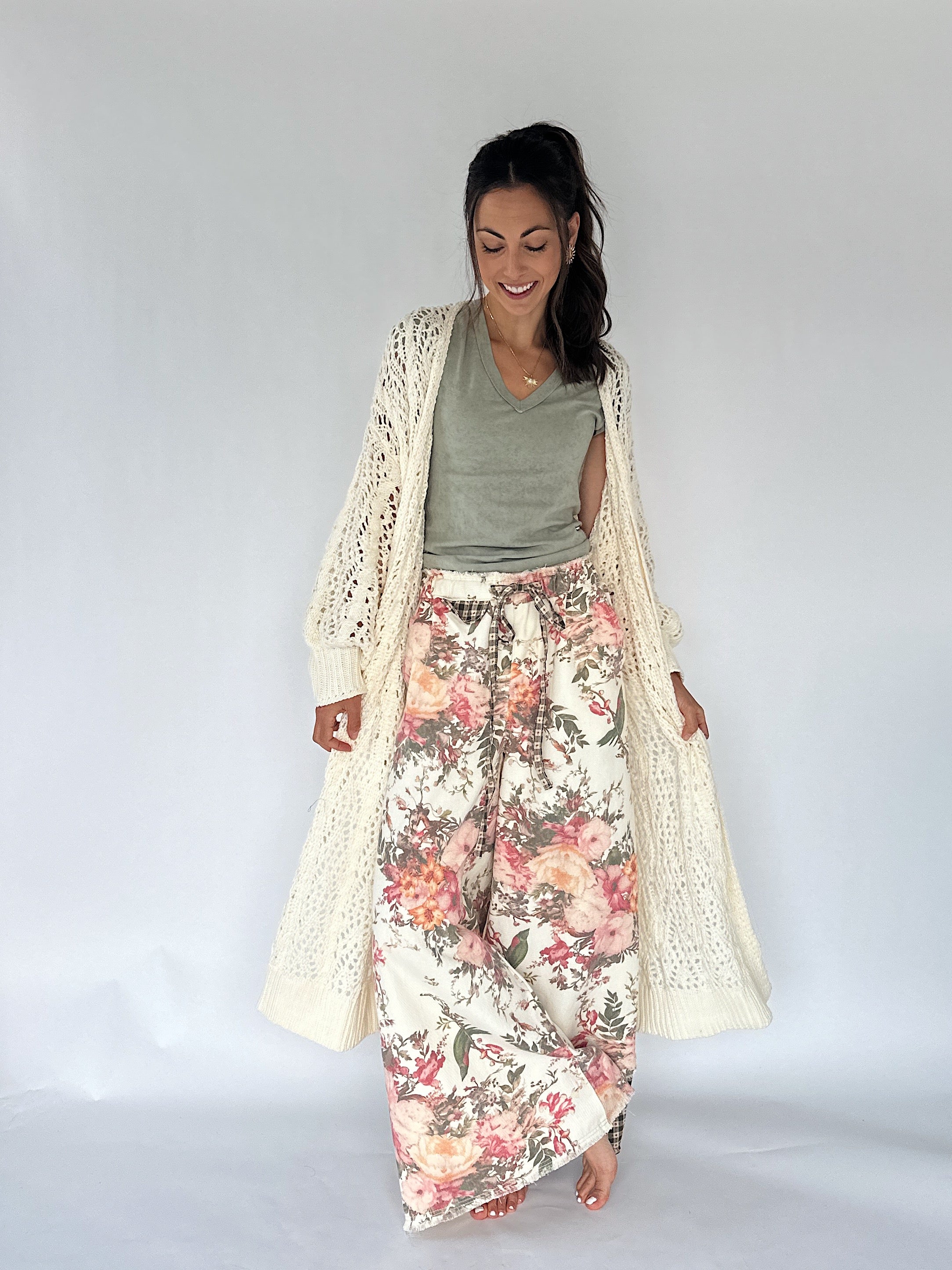 A woman modeling a bohemian-style outfit consisting of a cream open-front crochet cardigan, a sage green V-neck top, and wide-leg floral print pants with pink, red, and green flowers on a cream background.