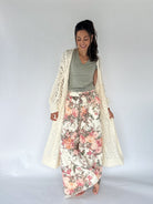 A woman modeling a bohemian-style outfit consisting of a cream open-front crochet cardigan, a sage green V-neck top, and wide-leg floral print pants with pink, red, and green flowers on a cream background.