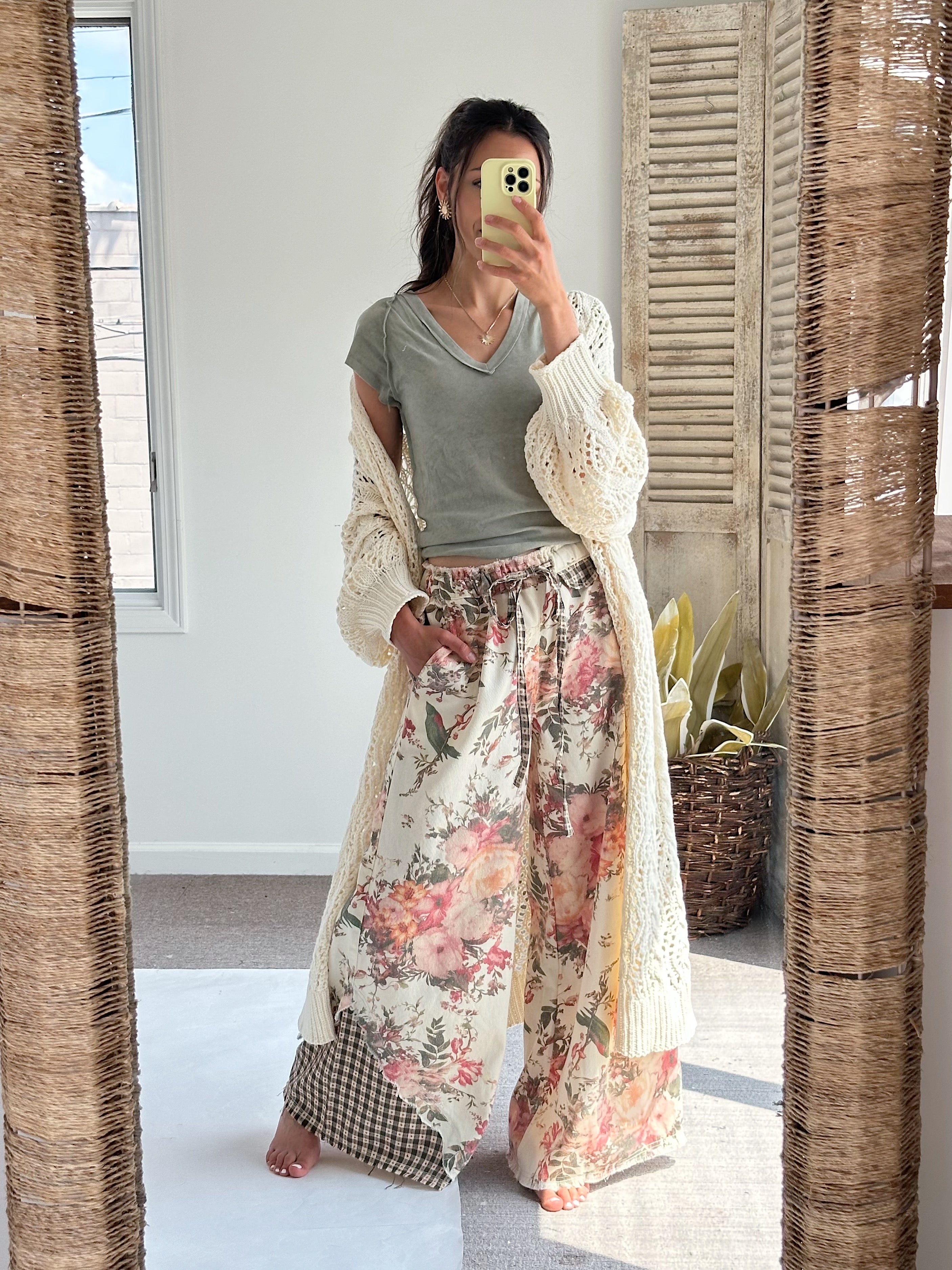 A woman takes a mirror selfie wearing a bohemian-style outfit consisting of a sage green V-neck t-shirt, a cream open-knit cardigan, and wide-leg floral print pants with a gingham leg overlay.