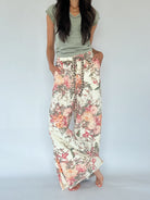 A woman wearing a sage green t-shirt and wide-leg floral pants with a pink, orange, and green botanical print on a cream background, standing against a light gray studio backdrop.