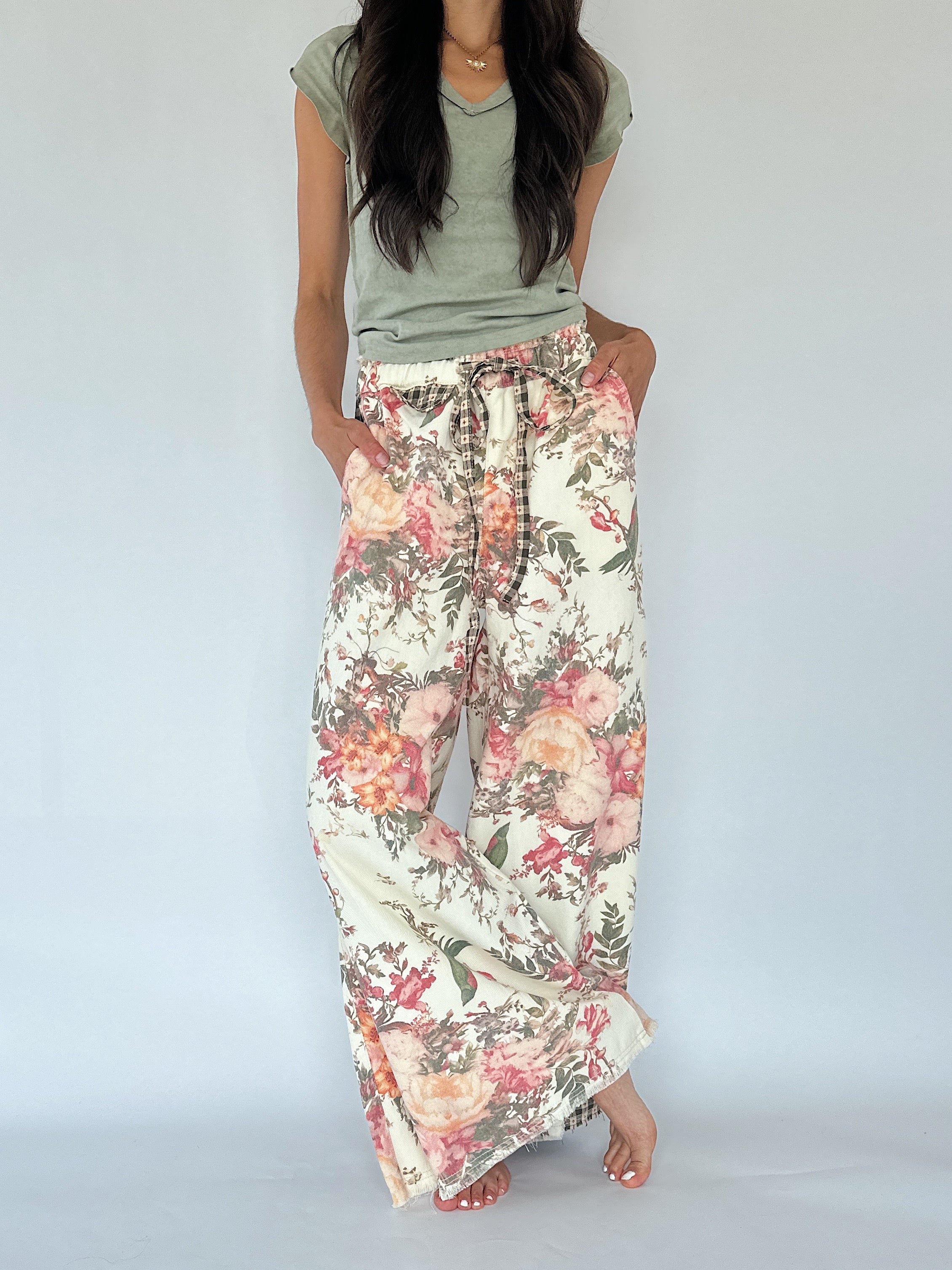 A woman wearing a sage green t-shirt and wide-leg floral pants with a pink, orange, and green botanical print on a cream background, standing against a light gray studio backdrop.