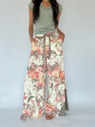 A model wearing wide-leg floral print pants with a drawstring waist and a green-and-white gingham side panel, paired with a sage green V-neck t-shirt against a plain light gray background.