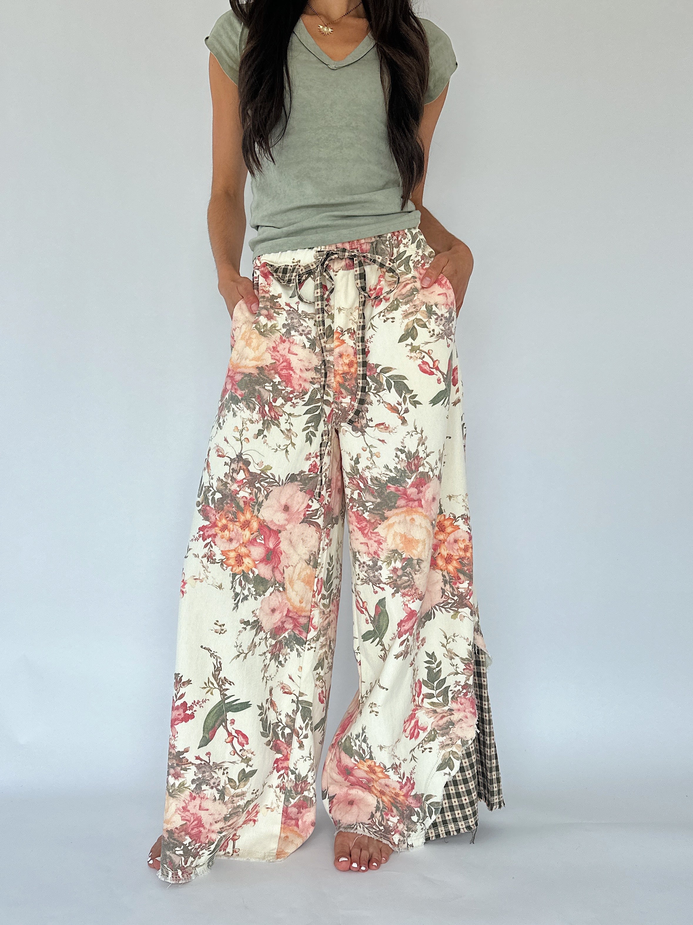 A model wearing wide-leg floral print pants with a drawstring waist and a green-and-white gingham side panel, paired with a sage green V-neck t-shirt against a plain light gray background.