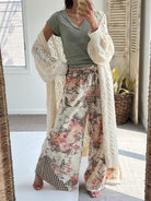 A woman takes a mirror selfie wearing a sage green t-shirt, a cream open-knit cardigan, and wide-leg floral pants with a gingham hem patch. The setting is a bright room with a window, shutters, and a potted plant.