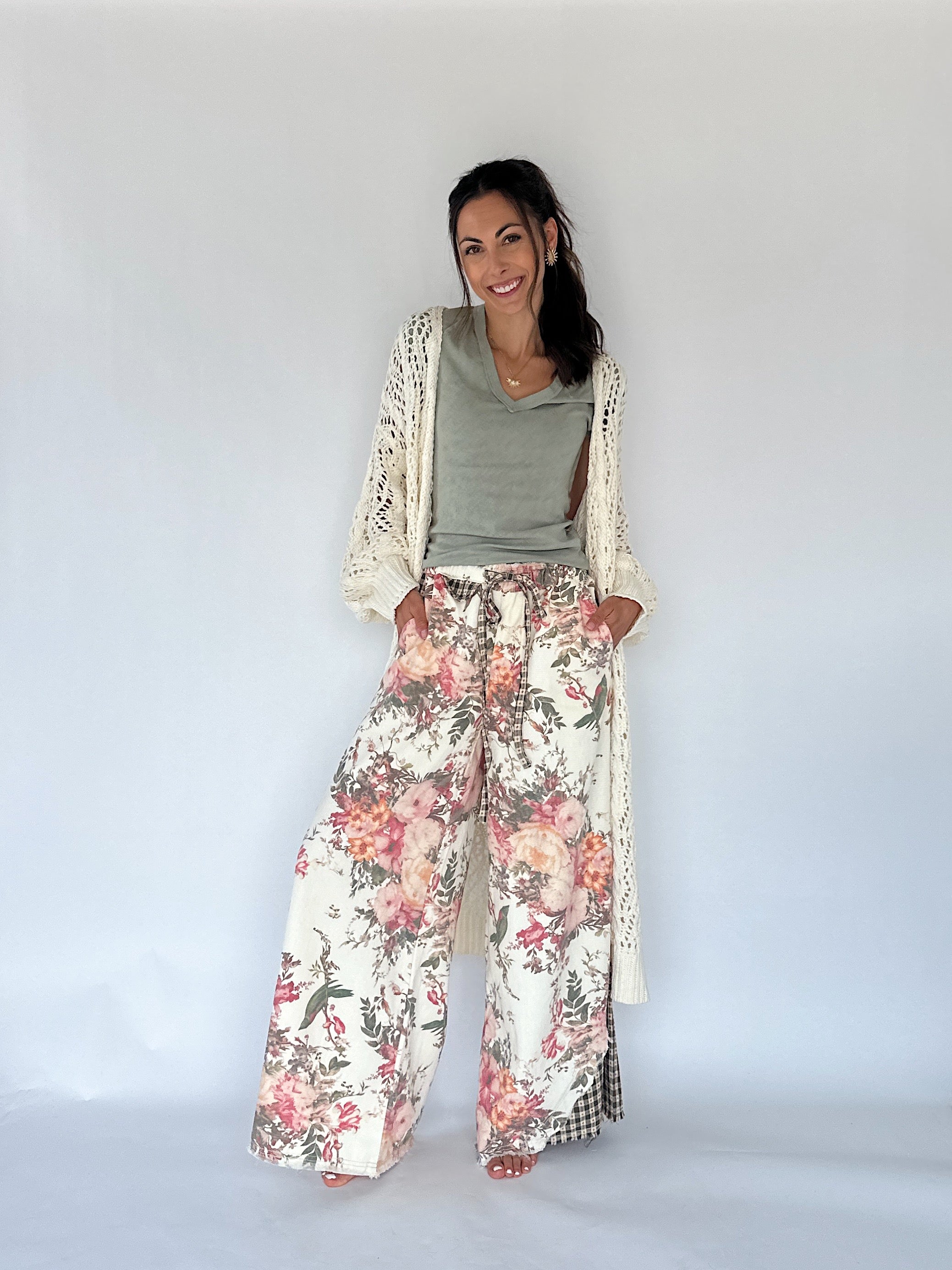 A woman modeling a bohemian-style outfit consisting of wide-leg floral print pants, a sage green top, and a cream open-knit cardigan, set against a plain white studio background.