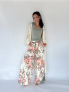 A woman modeling a bohemian-style outfit consisting of wide-leg floral print pants, a sage green top, and a cream open-knit cardigan, set against a plain white studio background.