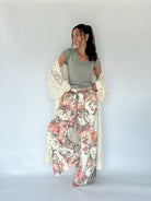 A woman modeling a bohemian-style outfit consisting of a sage green top, wide-leg floral print pants, and a cream crochet cardigan, set against a plain light gray studio background.