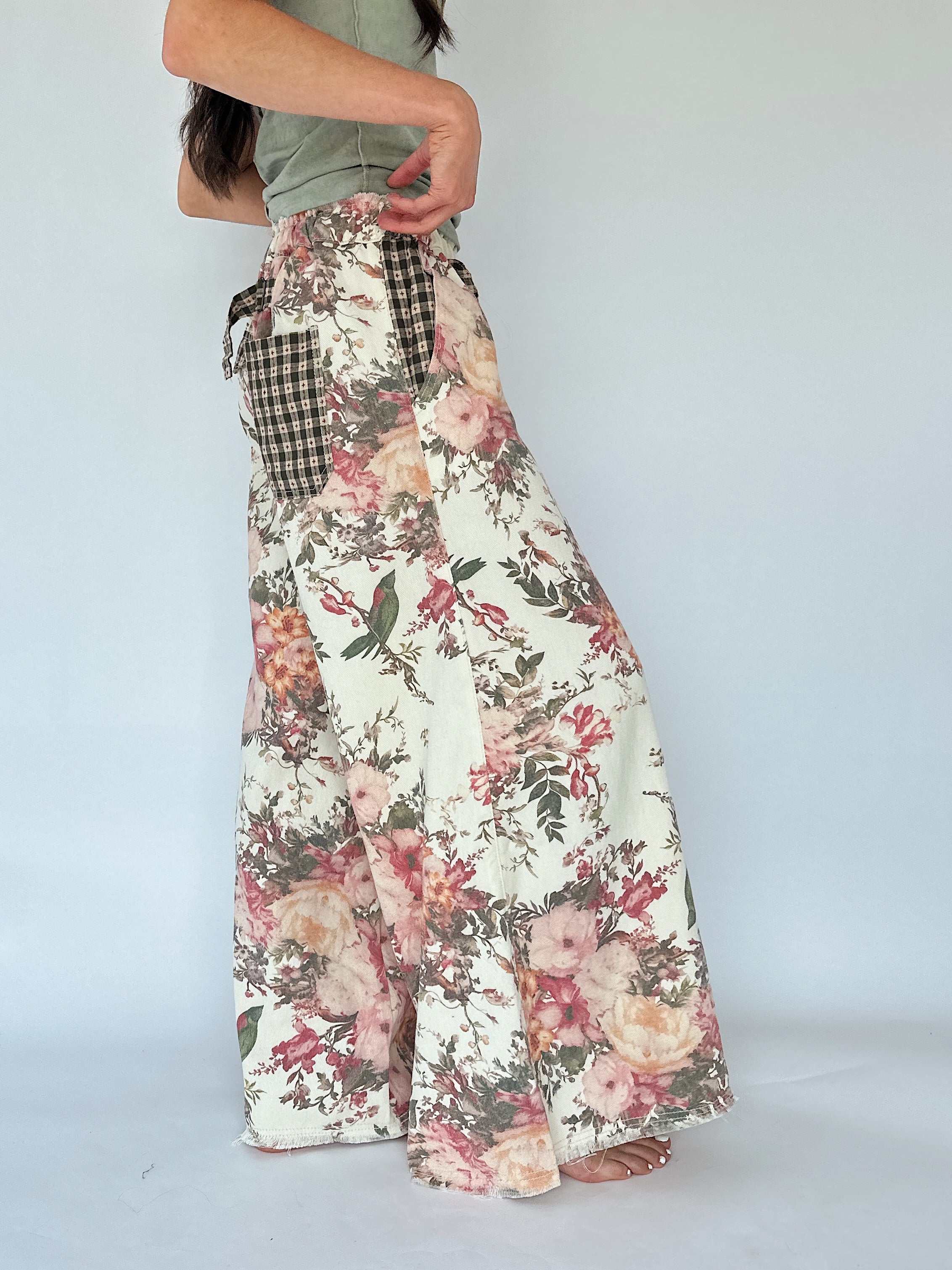 A person from the side wearing a pair of wide-leg floral pants with a gingham tie detail at the waist, shown from the torso down against a plain light gray studio background.