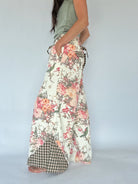 A woman wearing  long, wide-leg pants with a floral print on a cream background, featuring a green and white gingham checkered hem and side pocket detail, paired with a sage green sleeveless top against a light gray studio background.