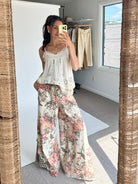 A woman takes a mirror selfie in a room, wearing a white sleeveless lace-trim camisole top and wide-leg floral pants with pink, green, and beige tones.
