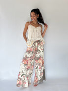 A woman modeling a bohemian-style two-piece outfit featuring a cream lace-trimmed camisole top and matching wide-leg pants with a pink, red, and green floral print, set against a plain white studio background.