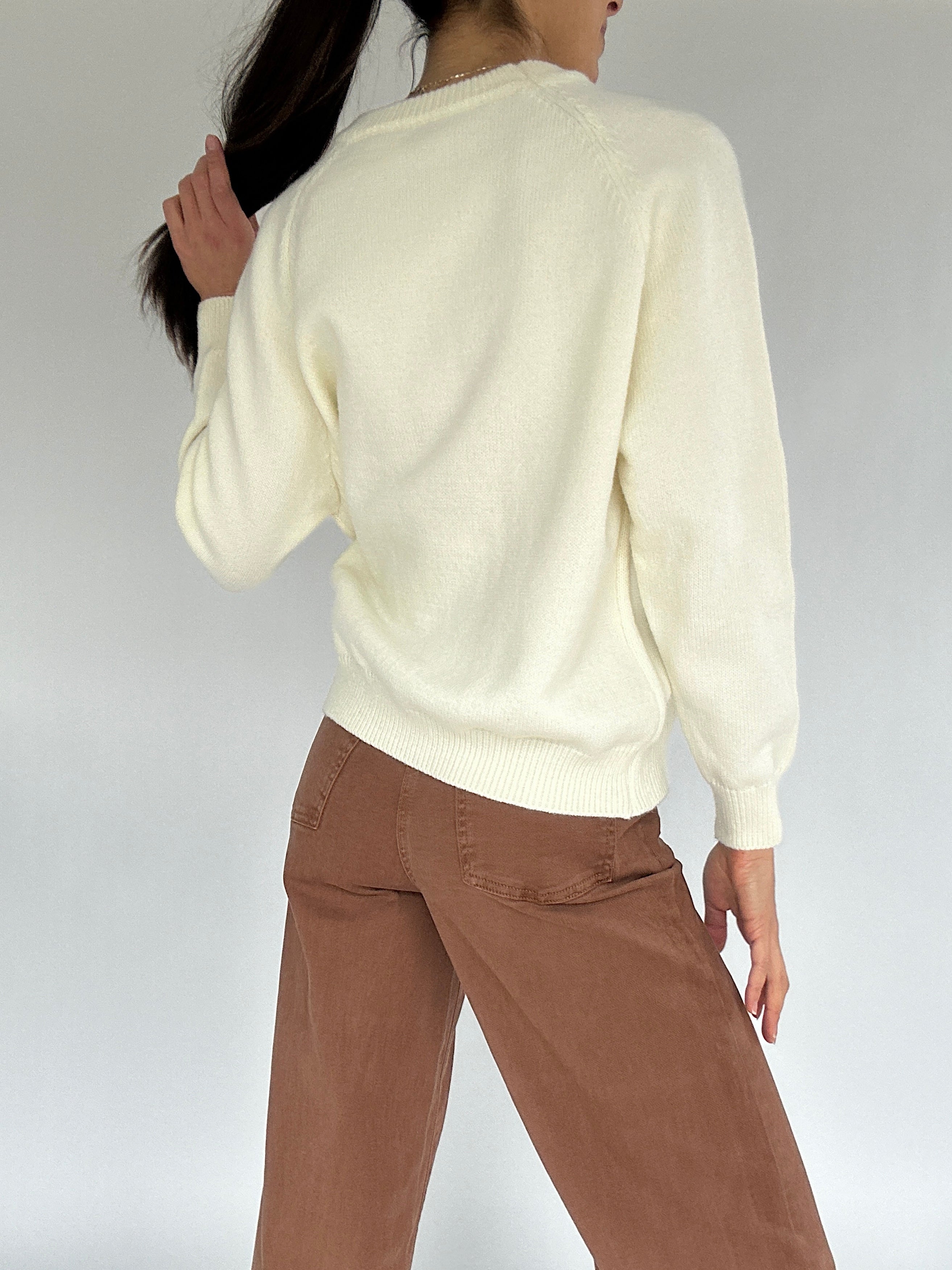Person wearing a cream sweater and brown pants against a white background showing back