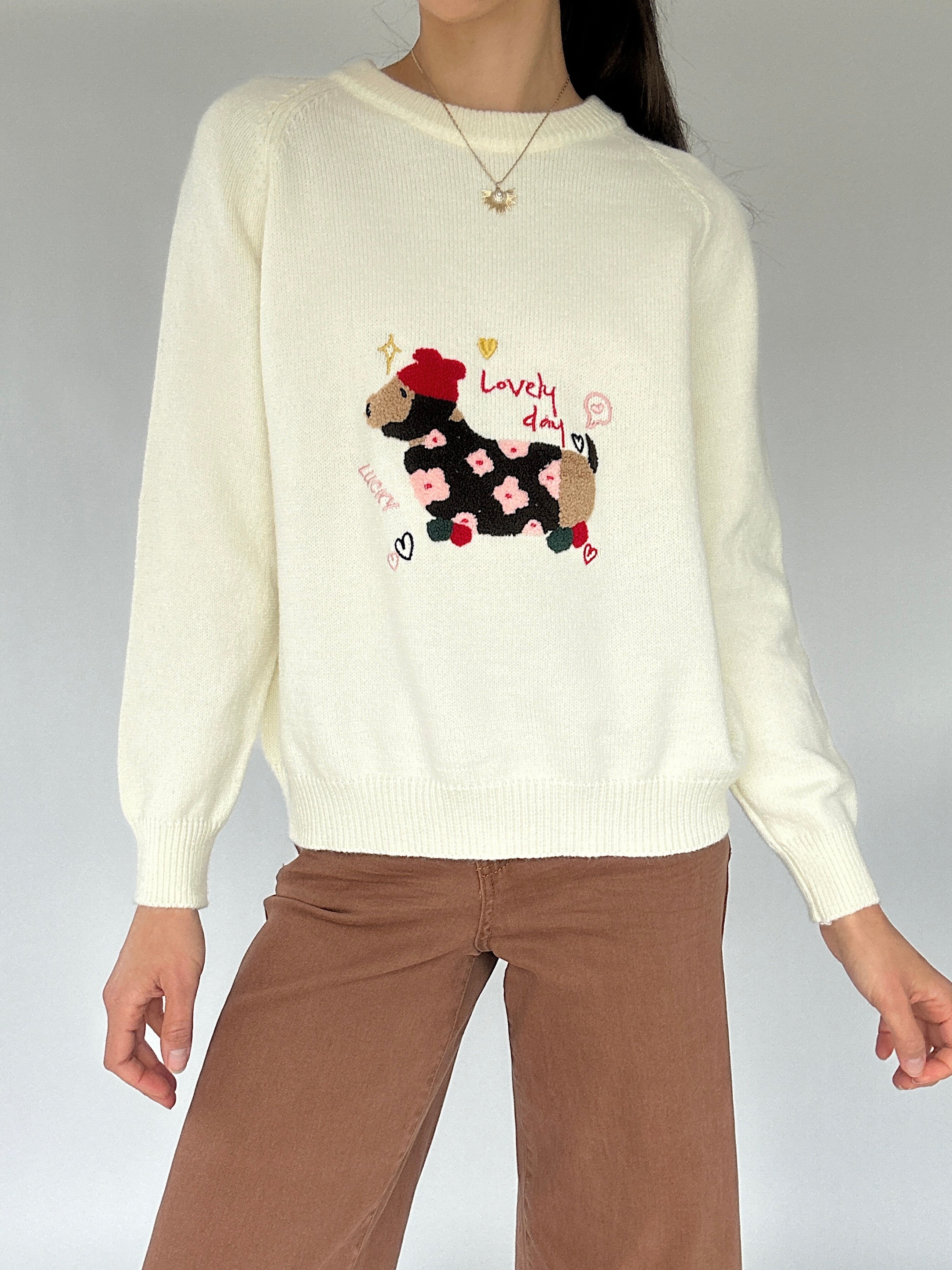 Person wearing a cream sweater with a dog design that says 'lovely day' and brown pants on a gray background