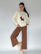 Woman wearing a cream sweater with a graphic design and brown pants on a white background