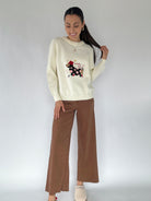 Woman wearing a cream sweater with a graphic dog design and brown pants on a white background