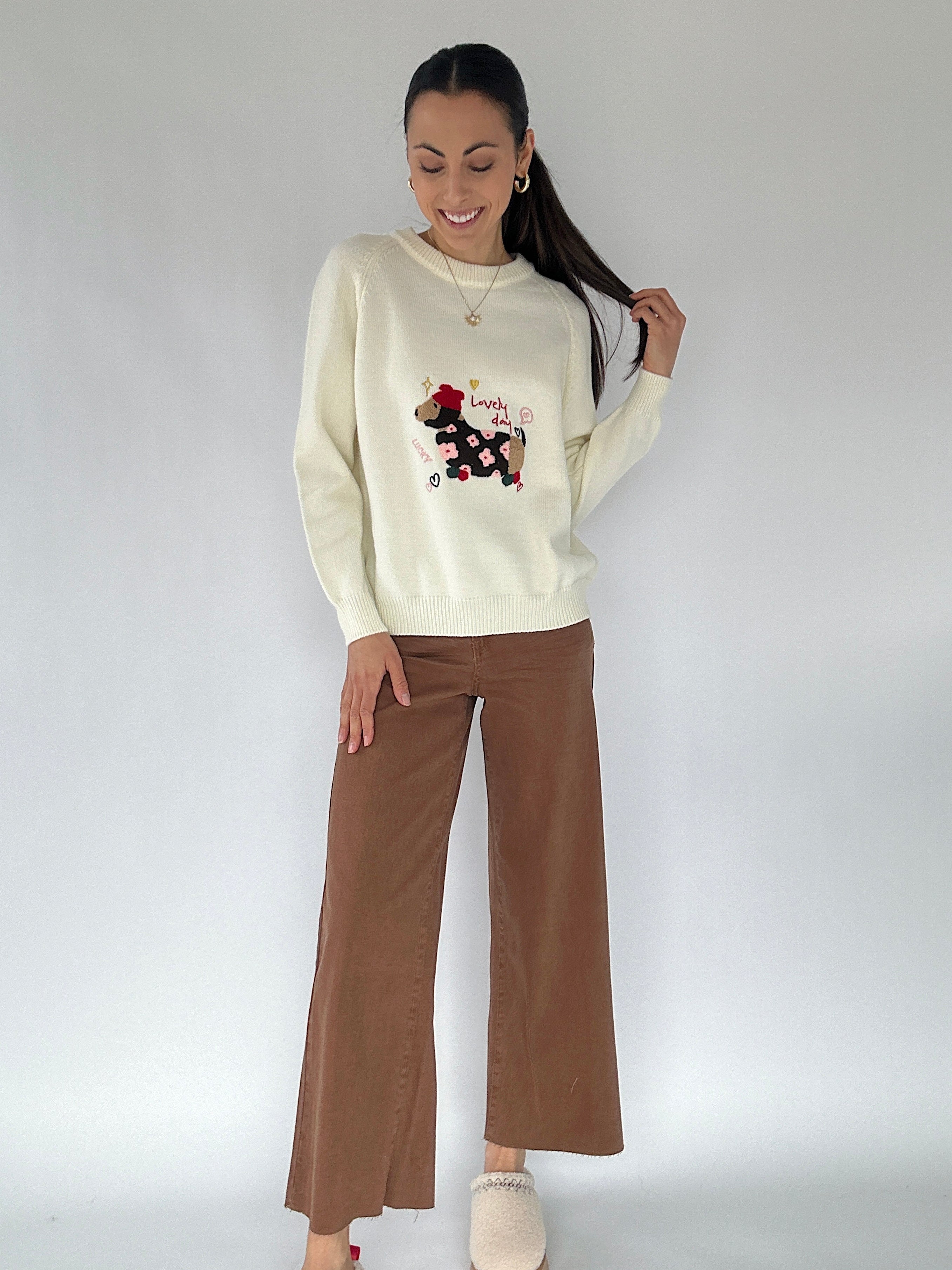 Woman wearing a cream sweater with a graphic dog design and brown pants on a white background