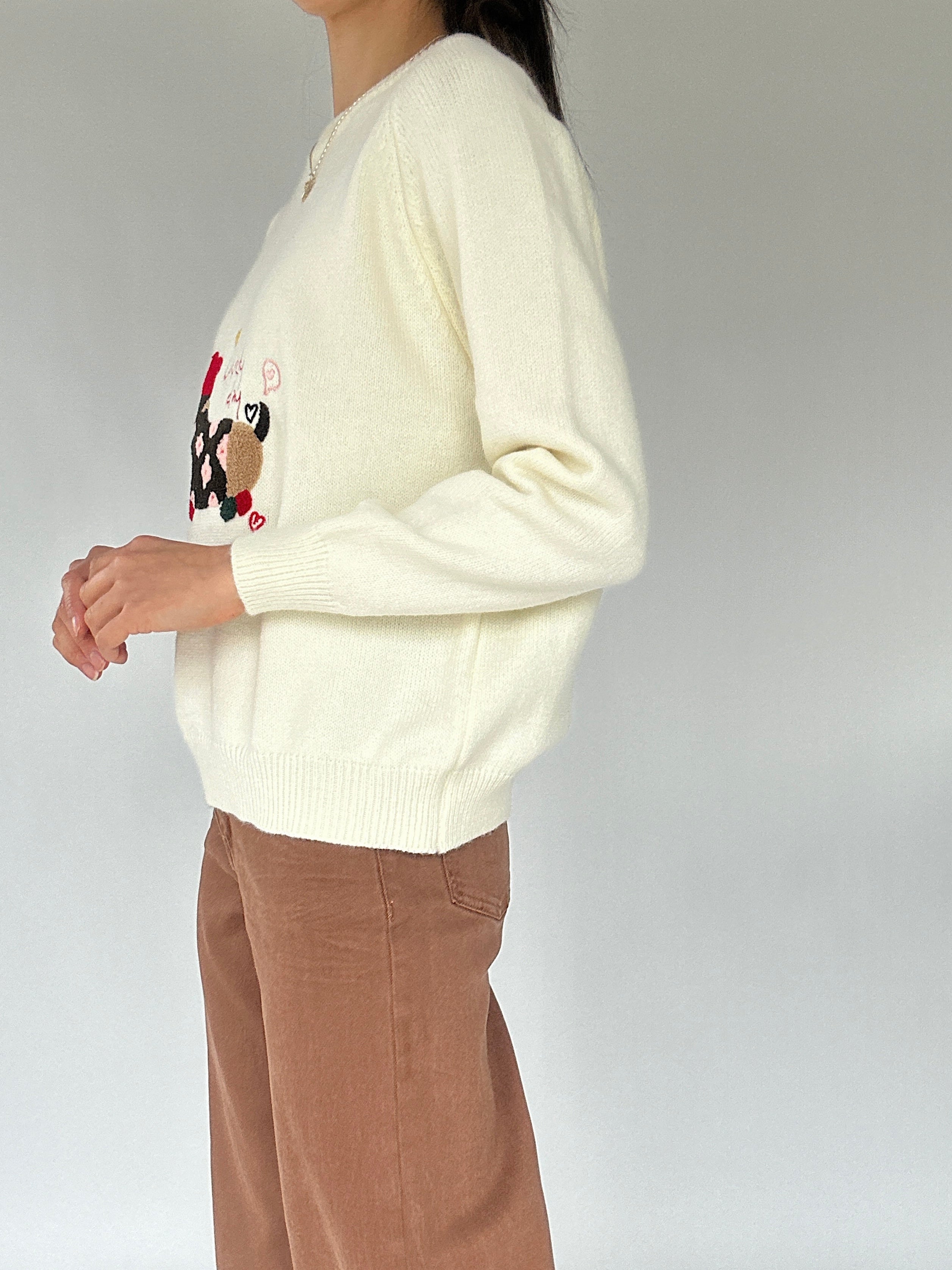 Person wearing a cream sweater with a design on the sleeve against a plain background