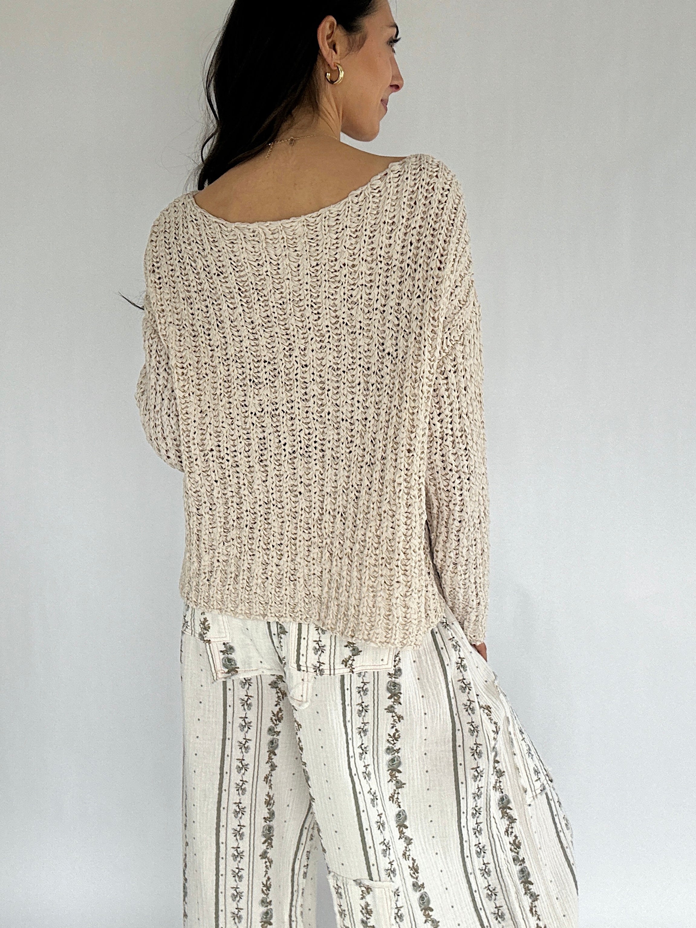 Woman wearing a beige knitted sweater and white patterned pants against a plain background from back