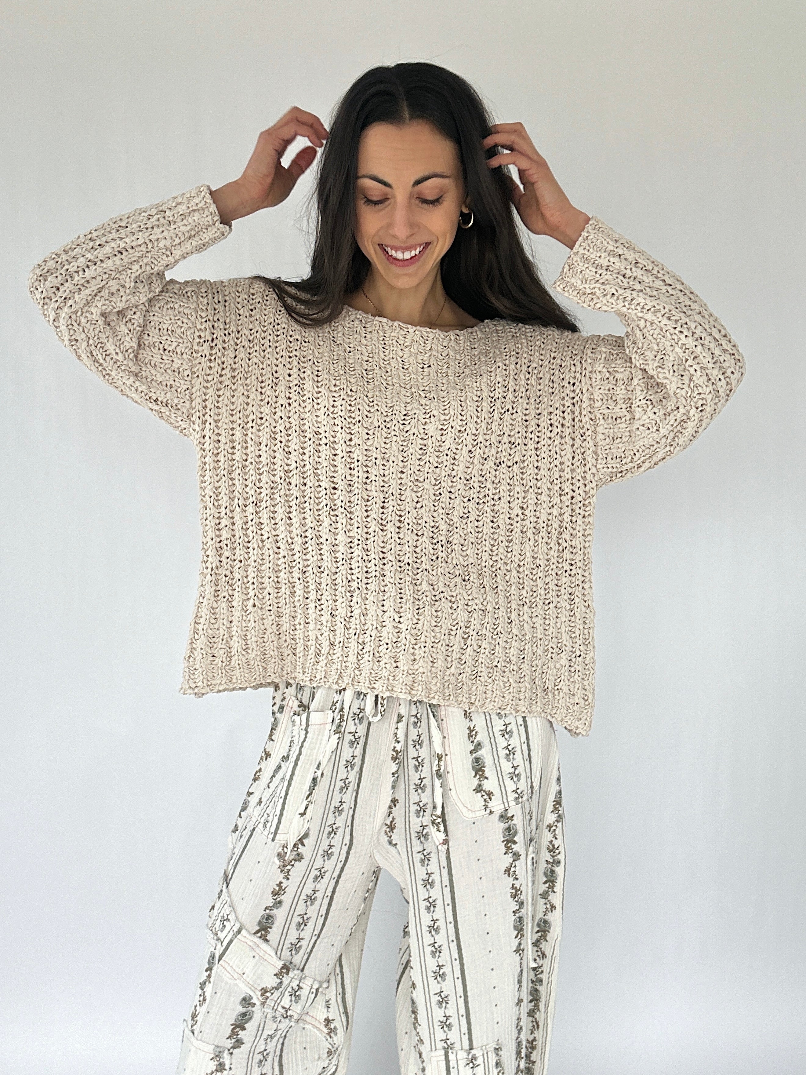 Woman wearing a beige knitted sweater and patterned pants against a white background