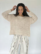 Woman wearing a beige knitted sweater and patterned pants against a white background