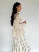 Woman wearing a beige knitted sweater and patterned pants against a white background from side