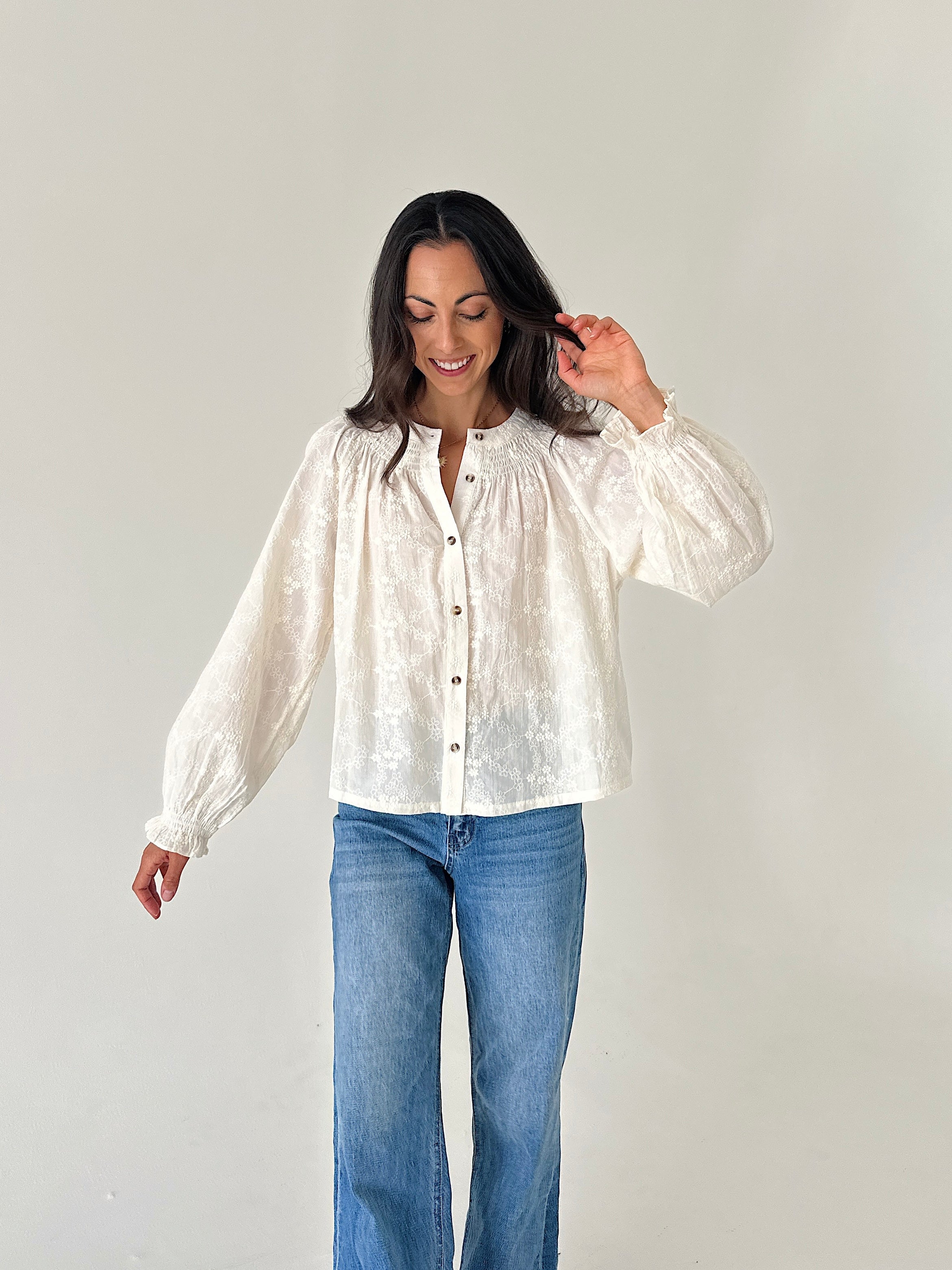 A woman modeling a white textured long-sleeve blouse with a button-down front, paired with blue jeans, against a plain light gray background.