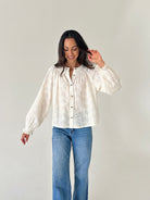 A woman modeling a white textured long-sleeve blouse with a button-down front, paired with blue jeans, against a plain light gray background.