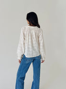 A woman viewed from behind wearing a white long-sleeve blouse with delicate floral embroidery and blue jeans, standing against a plain light gray wall.