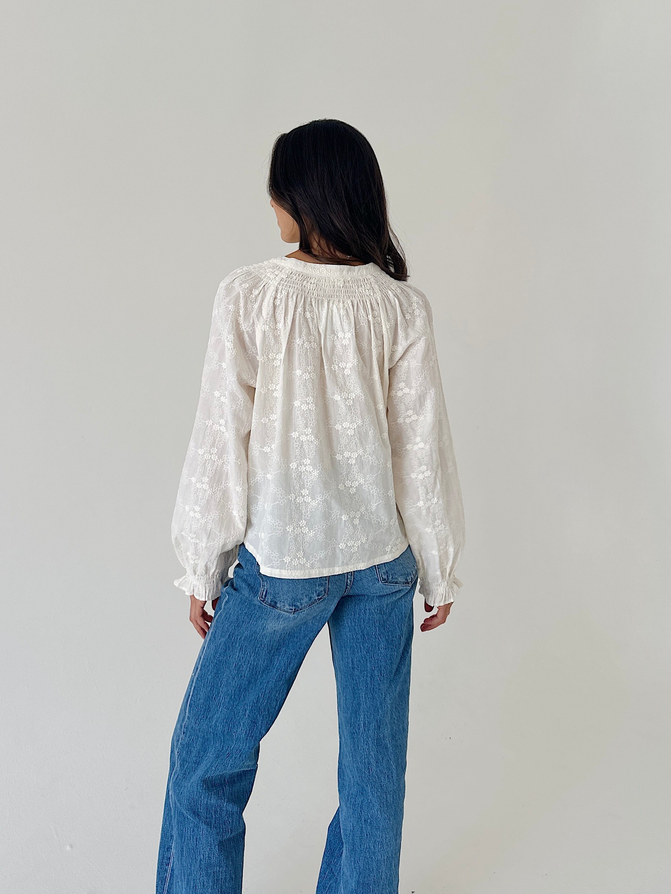A woman viewed from behind wearing a white long-sleeve blouse with delicate floral embroidery and blue jeans, standing against a plain light gray wall.