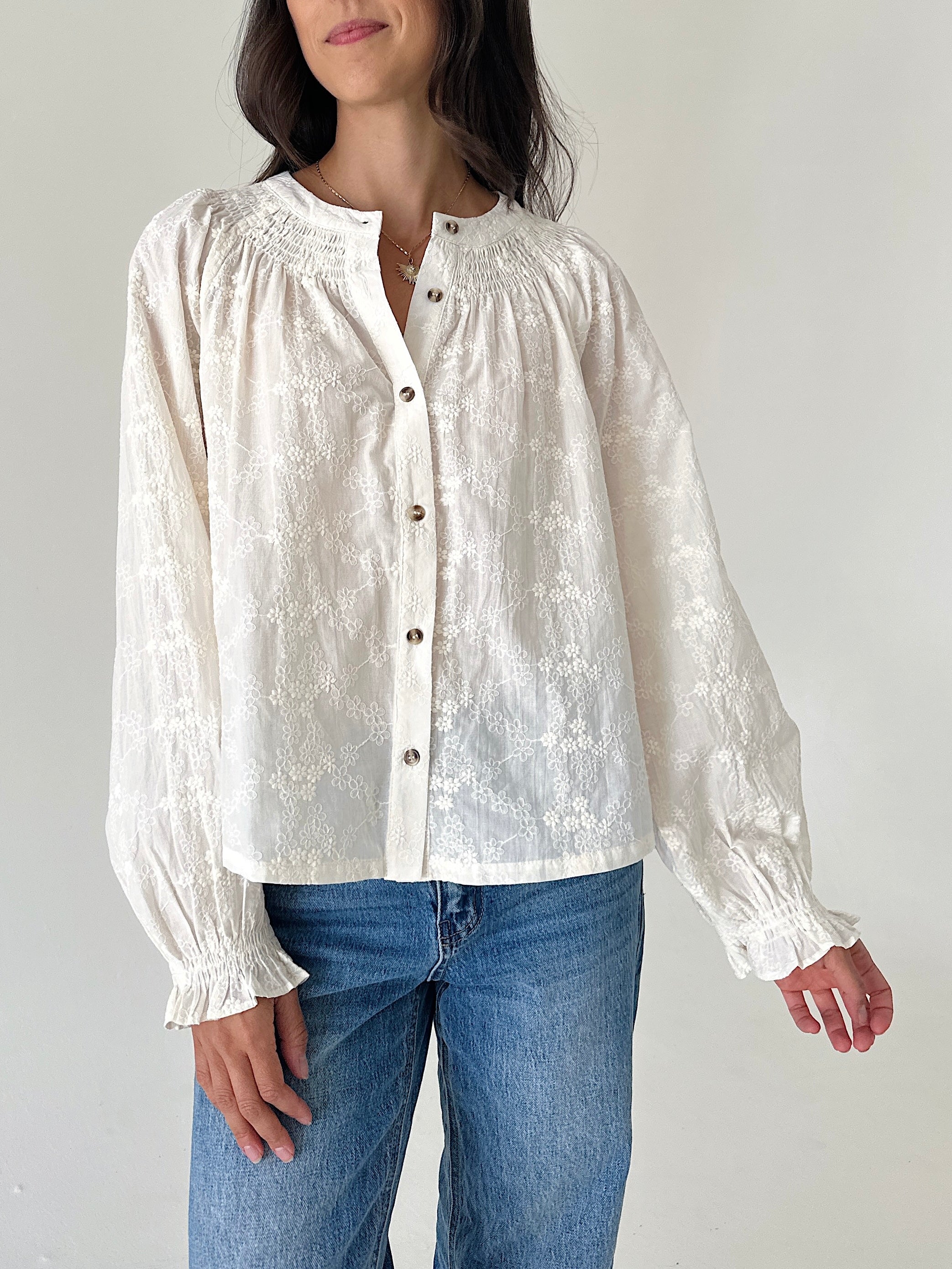 A woman wearing a cream-colored, long-sleeve lace blouse with gathered cuffs and a button-down front, paired with blue jeans, against a plain light background.