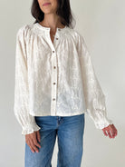 A woman wearing a cream-colored, long-sleeve lace blouse with gathered cuffs and a button-down front, paired with blue jeans, against a plain light background.