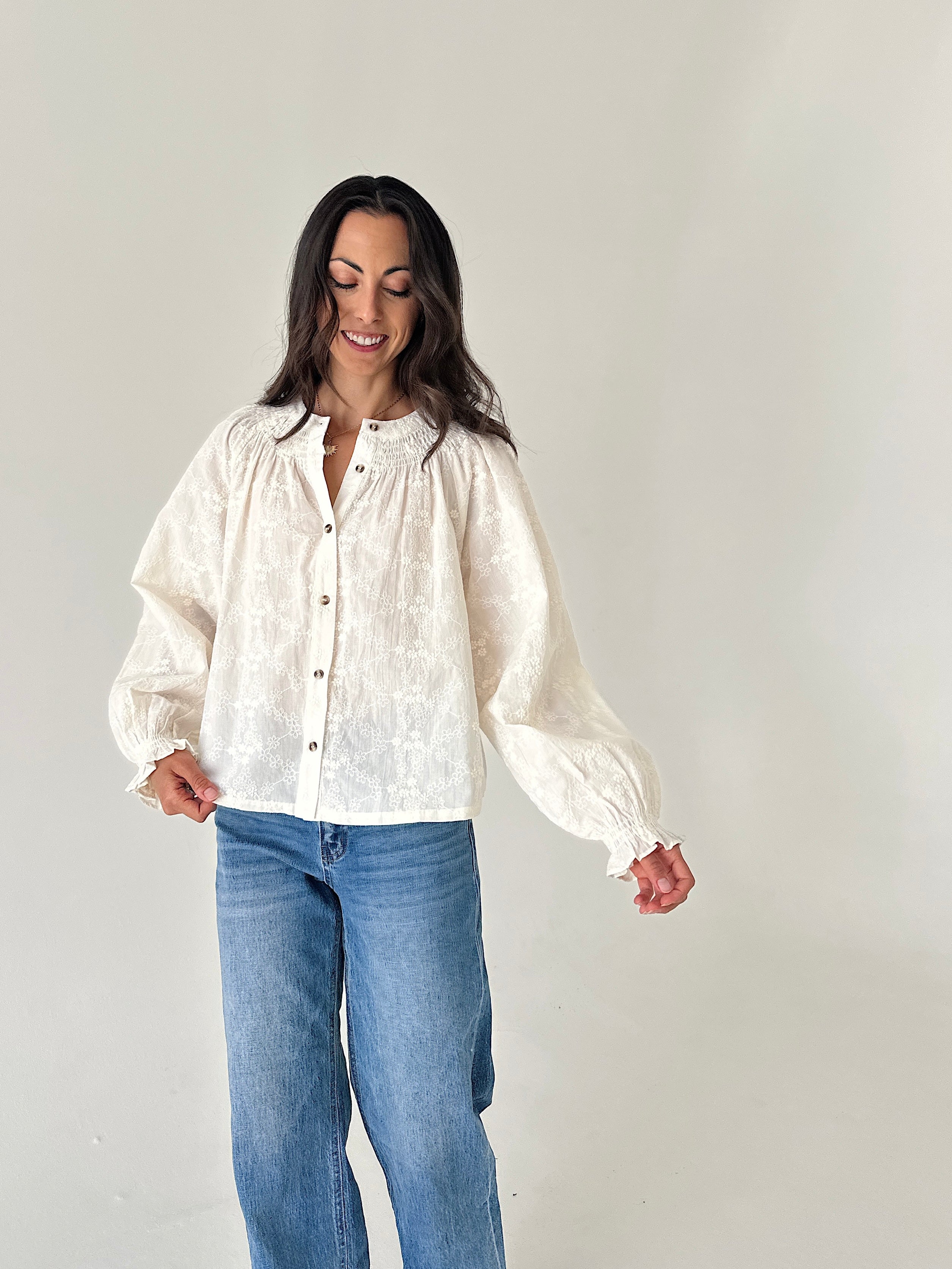 A woman wearing a cream-colored, textured long-sleeve blouse with gathered cuffs and a button-down front, paired with blue jeans, standing against a plain light gray background.